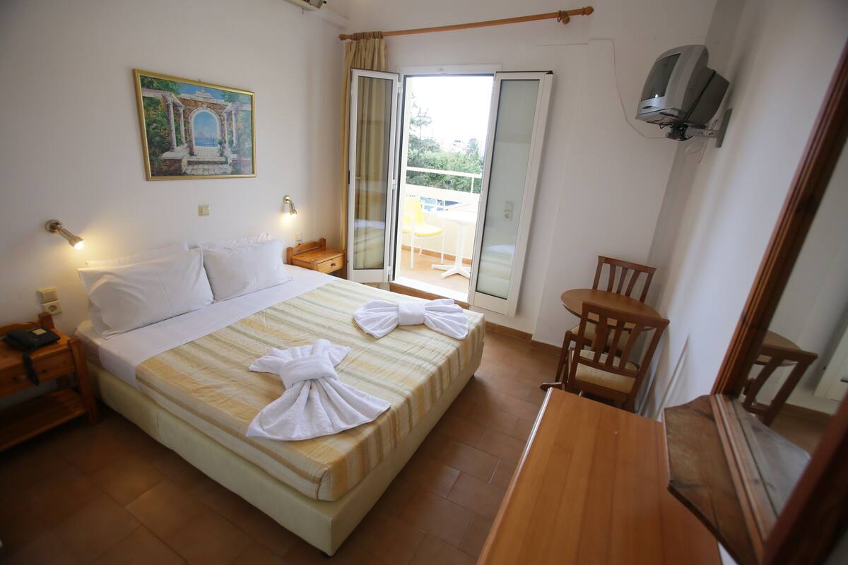 Ein Hotelzimmer mit Bett, Holzstuhl, Tisch, Fernseher und einem Balkon mit Sitzgelegenheit.