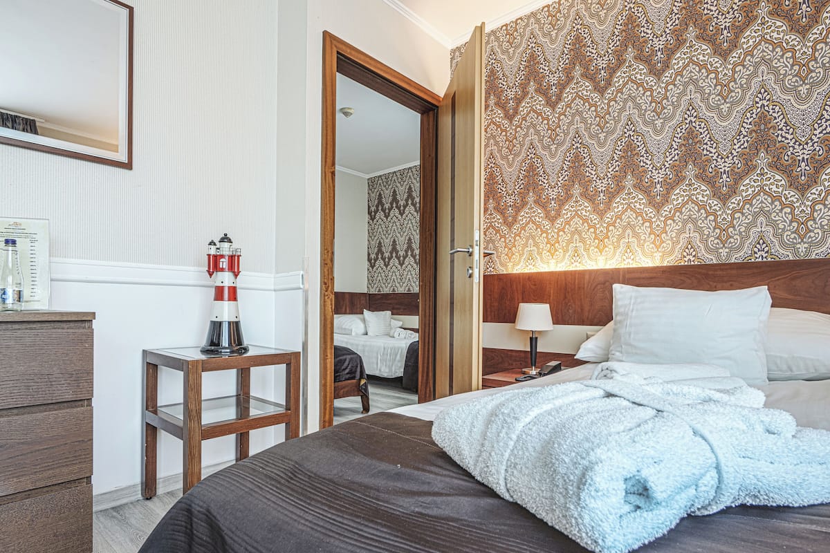 A hotel room with a large bed, a bedside table with a lamp, a small stool with a bottle, and a patterned wallpaper.