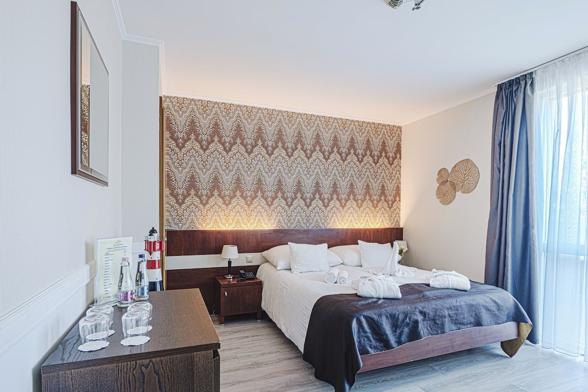 A hotel room with a large bed, a wooden nightstand, a mirror, and a patterned wallpaper.