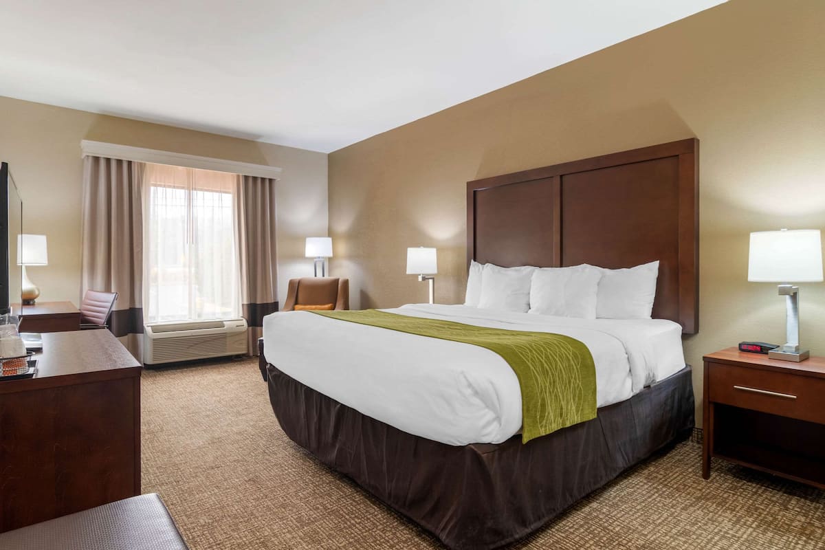 Una habitación de hotel con una cama grande, dos mesitas de noche con lámparas, un escritorio con silla, un ventanal con cortinas y un equipo de aire acondicionado.