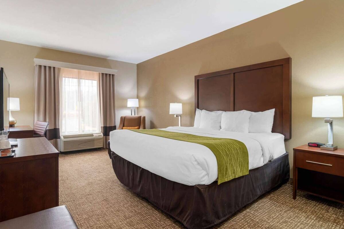 Una habitación de hotel con una cama grande, un escritorio, una silla, una lámpara y una ventana con cortinas.