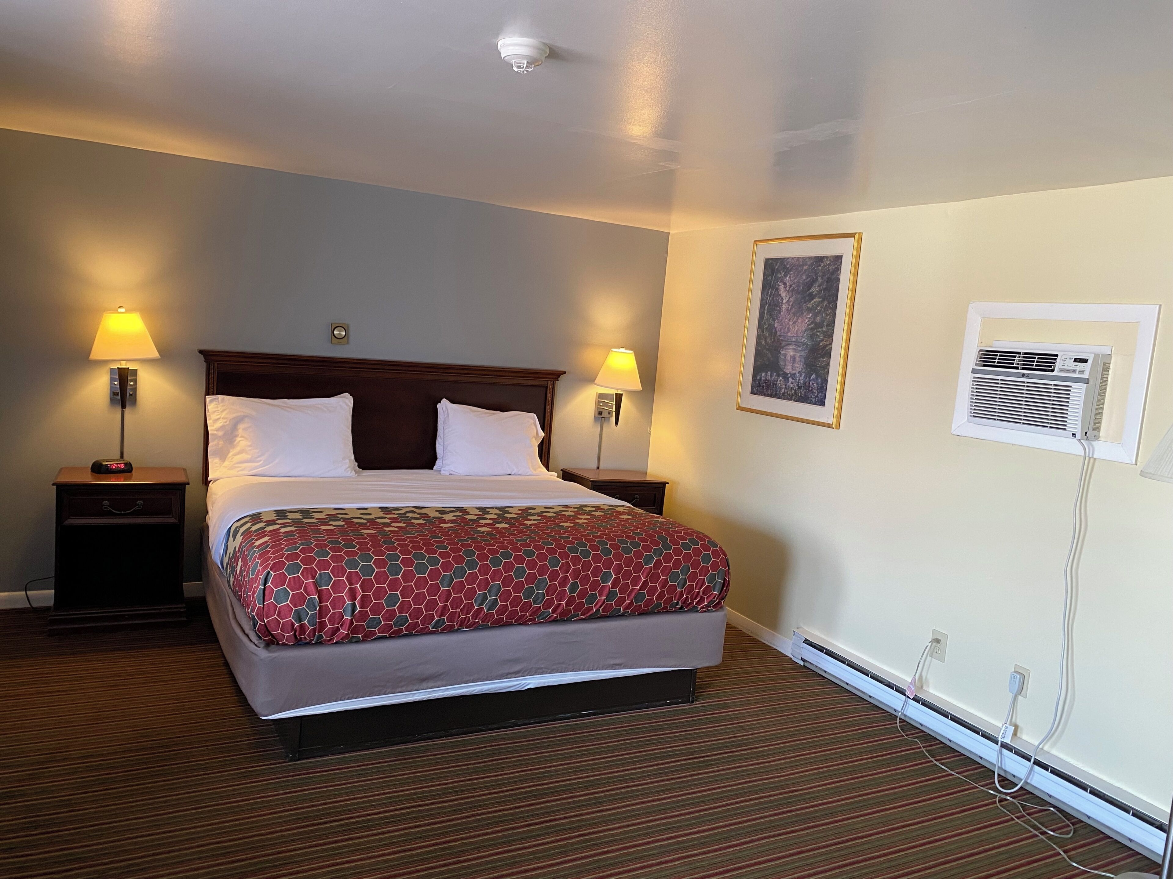 Habitación de hotel con cama, dos lámparas de noche, aire acondicionado montado en la pared y un cuadro enmarco.