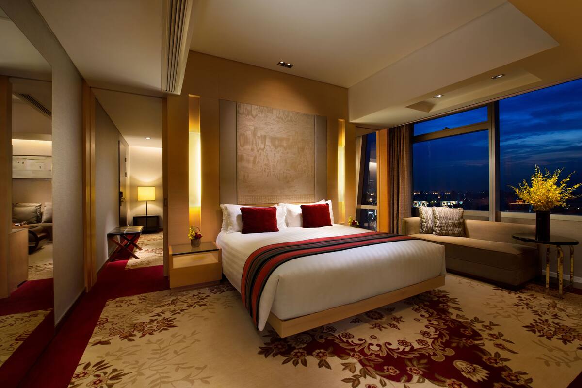 Une chambre d’hôtel moderne avec un grand lit, un canapé, un bureau et une vue sur la ville la nuit.