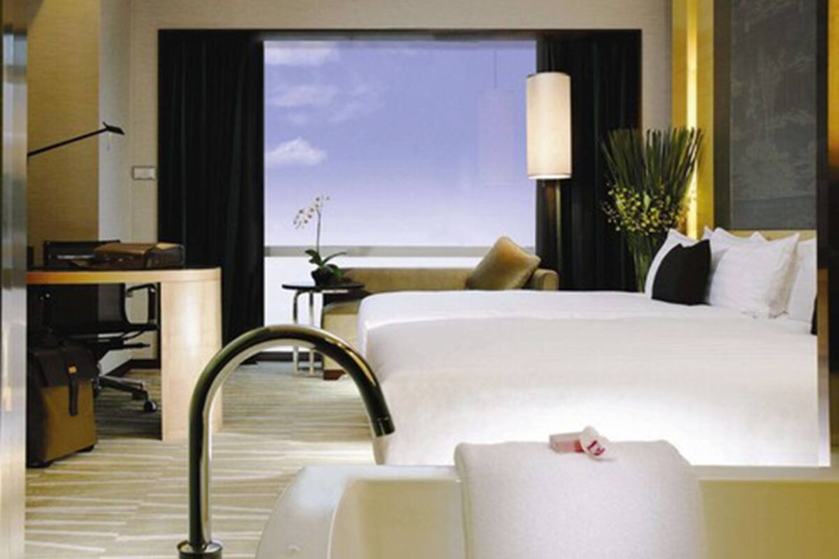 Une chambre d’hôtel avec un grand lit, un bureau avec un ordinateur, une chaise, une petite table avec une plante et une vue sur le ciel par la fenêtre.