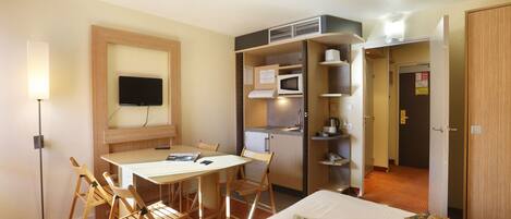 A hotel room with a bed, a desk with chairs, a television, a microwave, and a kitchenette.