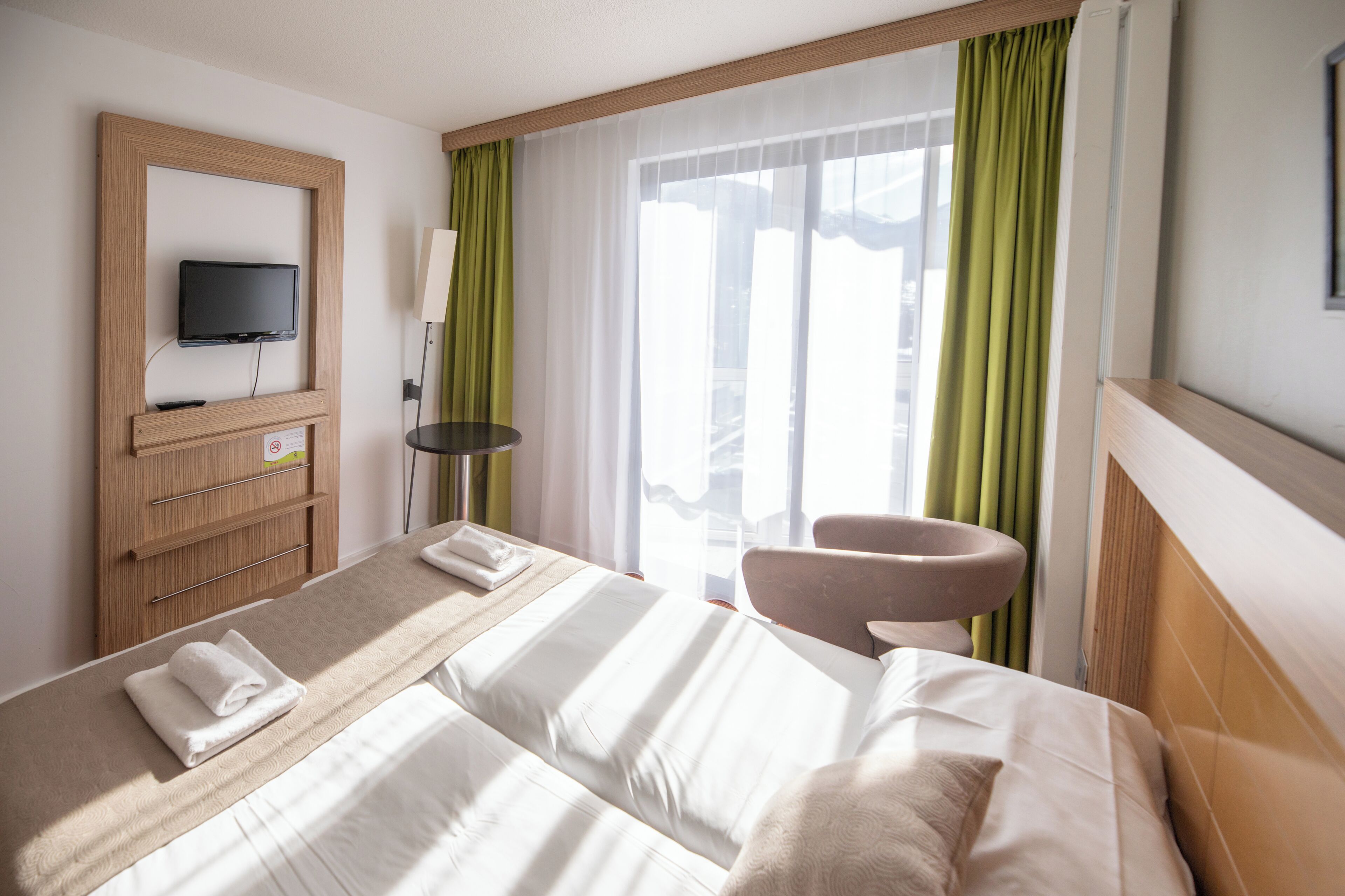A hotel room with a bed, a TV mounted on a wooden panel, a chair, and a window with green curtains.