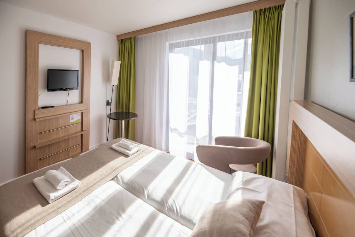 Ein Hotelzimmer mit einem Bett, einem an einer Holzplatte montierten Fernseher, einem Sessel und einem Fenster mit grünen Vorhängen.
