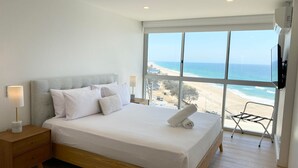 A bedroom with a large window overlooking a beach, a bed with white linens, a nightstand with a lamp, and a wooden floor.