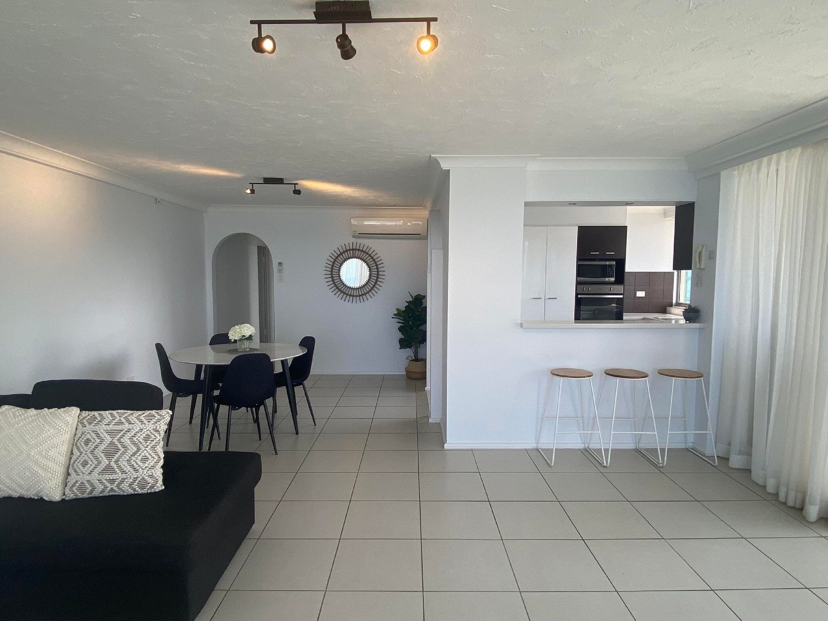A modern living room with a black sofa, a dining area with a round table, and a kitchen with a bar area.