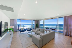 A modern living room with a large sofa, two armchairs, a coffee table, and a TV. The room has large windows offering a view of the ocean. - Hi Surf Beachfront Resort Apartments (Surfers Paradise)