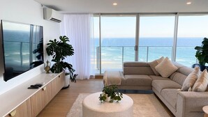 A modern living room with a large TV, a beige sofa, a round coffee table, and a view of the ocean.