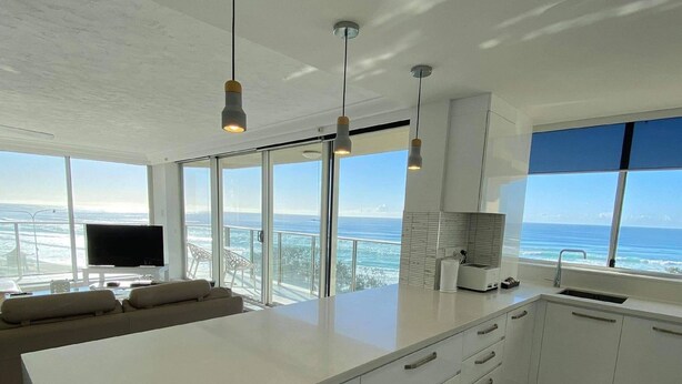 A modern kitchen with a large island, white cabinetry, and pendant lighting. A large window offers a view of the ocean.