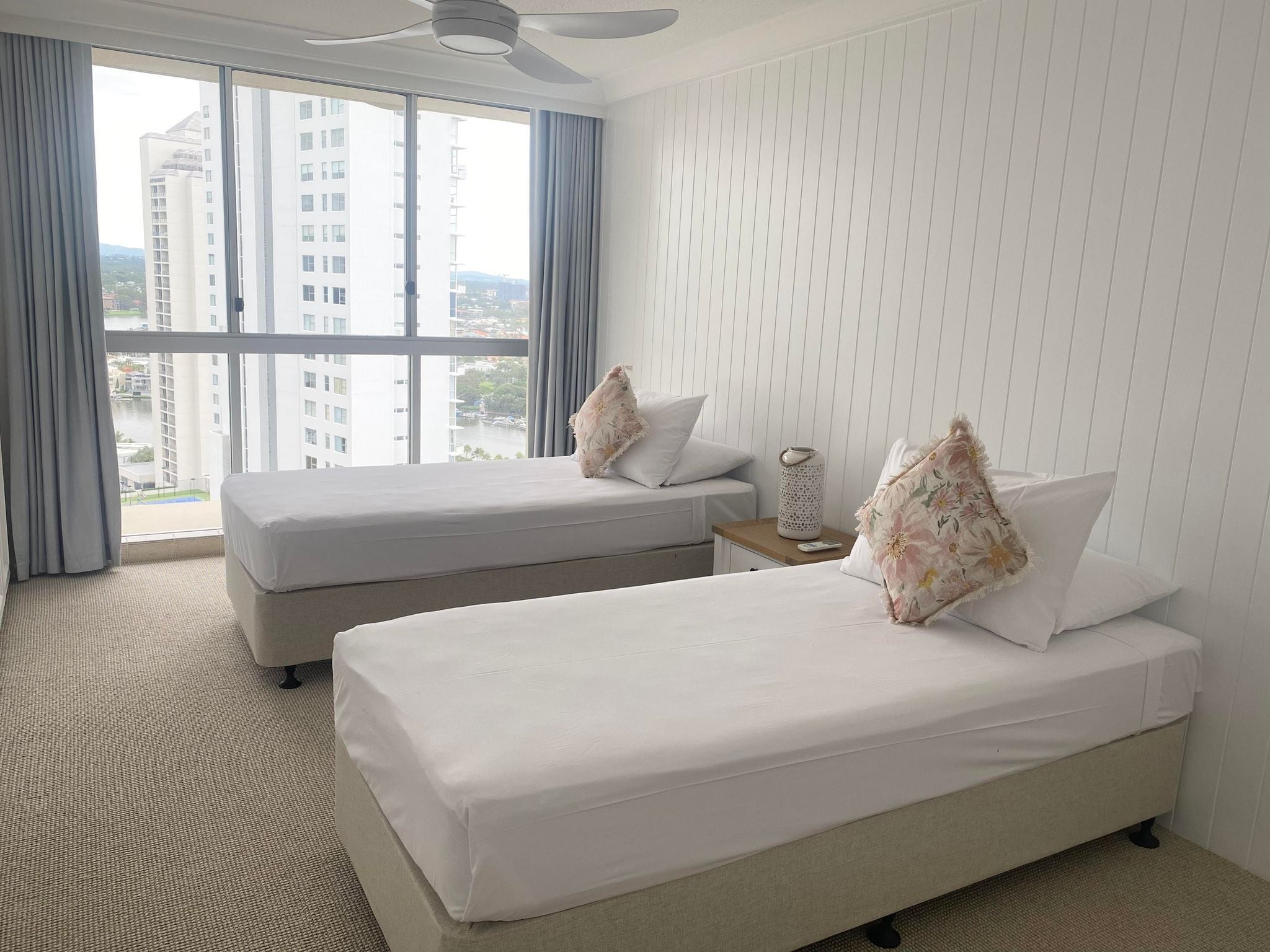 A hotel room with two beds, a large window with a view of buildings, a ceiling fan, and a small table with a vase.