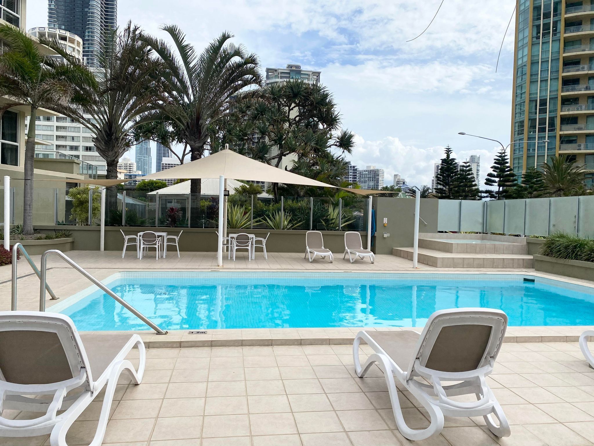 Une piscine entourée de palmiers et de bâtiments modernes, avec des chaises longues blanches et un auvent.