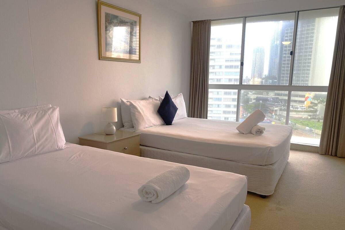 A hotel room with two beds, a nightstand, a lamp, and a large window with a city view.