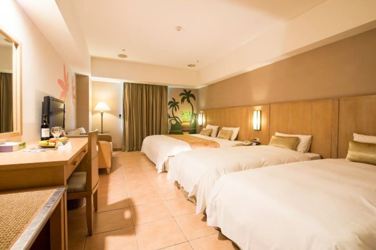 Habitación de hotel con dos camas, un escritorio con silla, televisor y un mural de palmeras en la pared.