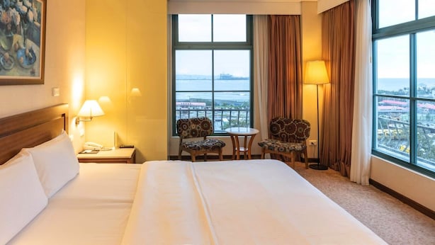 Hotelkamer met een groot bed, twee bedlampjes, een klein tafeltje met stoelen en uitzicht op een stadsgezicht door grote ramen.