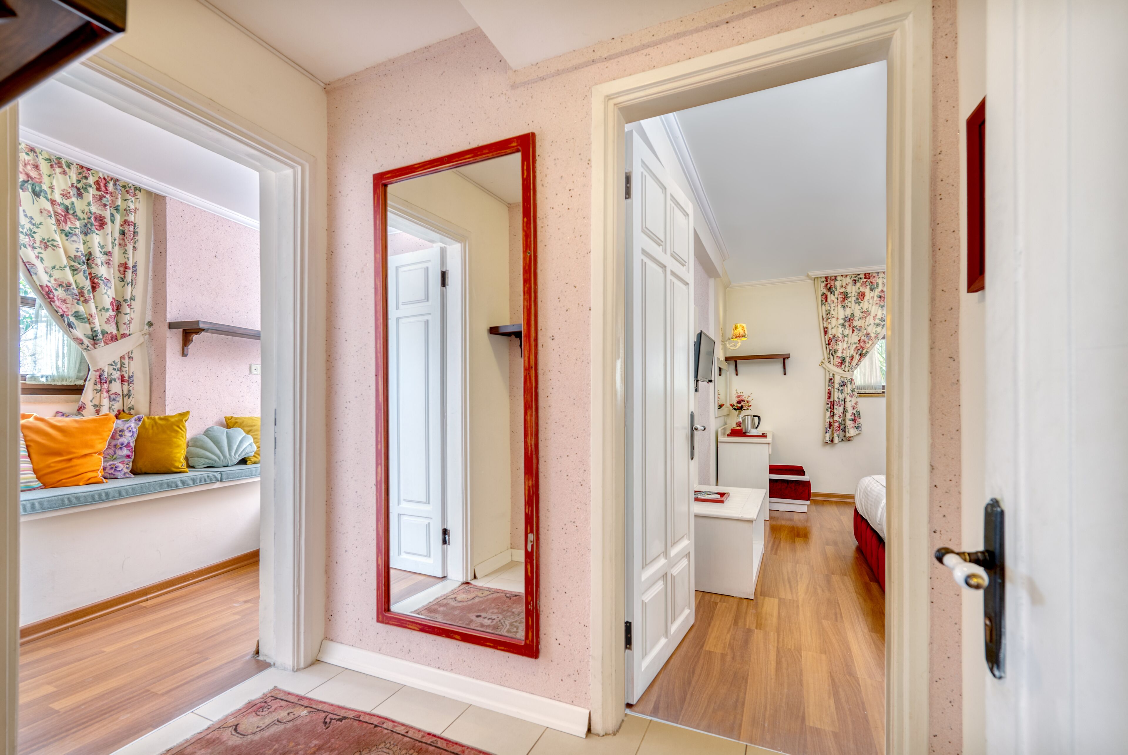 A hallway with a mirror, a bench with cushions, and a door leading to a bedroom with a bed and a nightstand.
