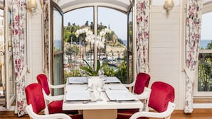 A dining area with a table set for four, surrounded by red chairs, with a view of a coastal landscape through large windows. - Adalya Port Hotel (Antalya)