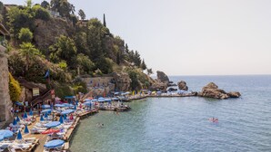 A coastal beach with sun loungers and umbrellas, clear water, and rocky cliffs. - Adalya Port Hotel (Antalya)