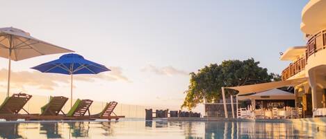 Zona de piscina con sillones de descanso y sombrillas, un bar y un árbol al fondo.