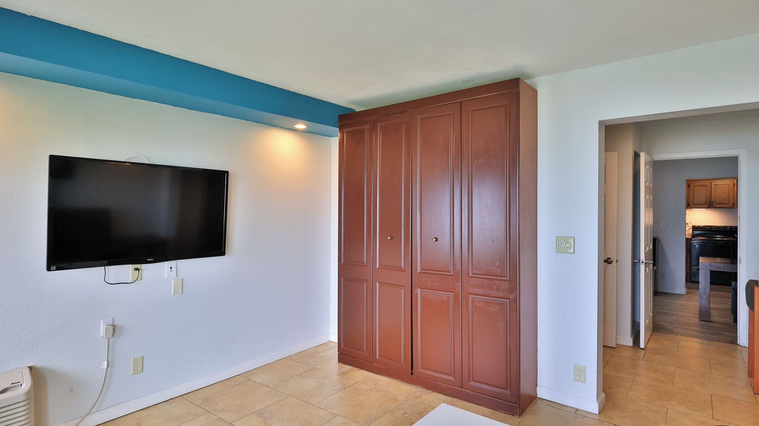Una habitación con televisor de pantalla plana en la pared, un armario de madera y una cocina a la vista a través de una puerta abierta.