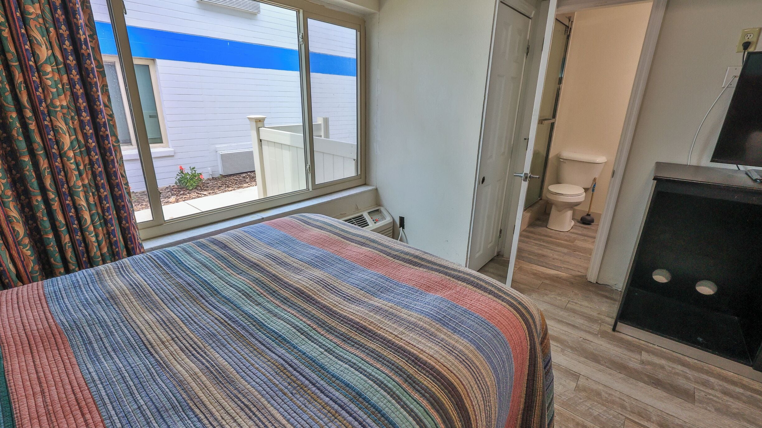 A hotel room with a large window, a bed with a striped bedspread, a small bathroom with a toilet and sink, and a TV on a stand.