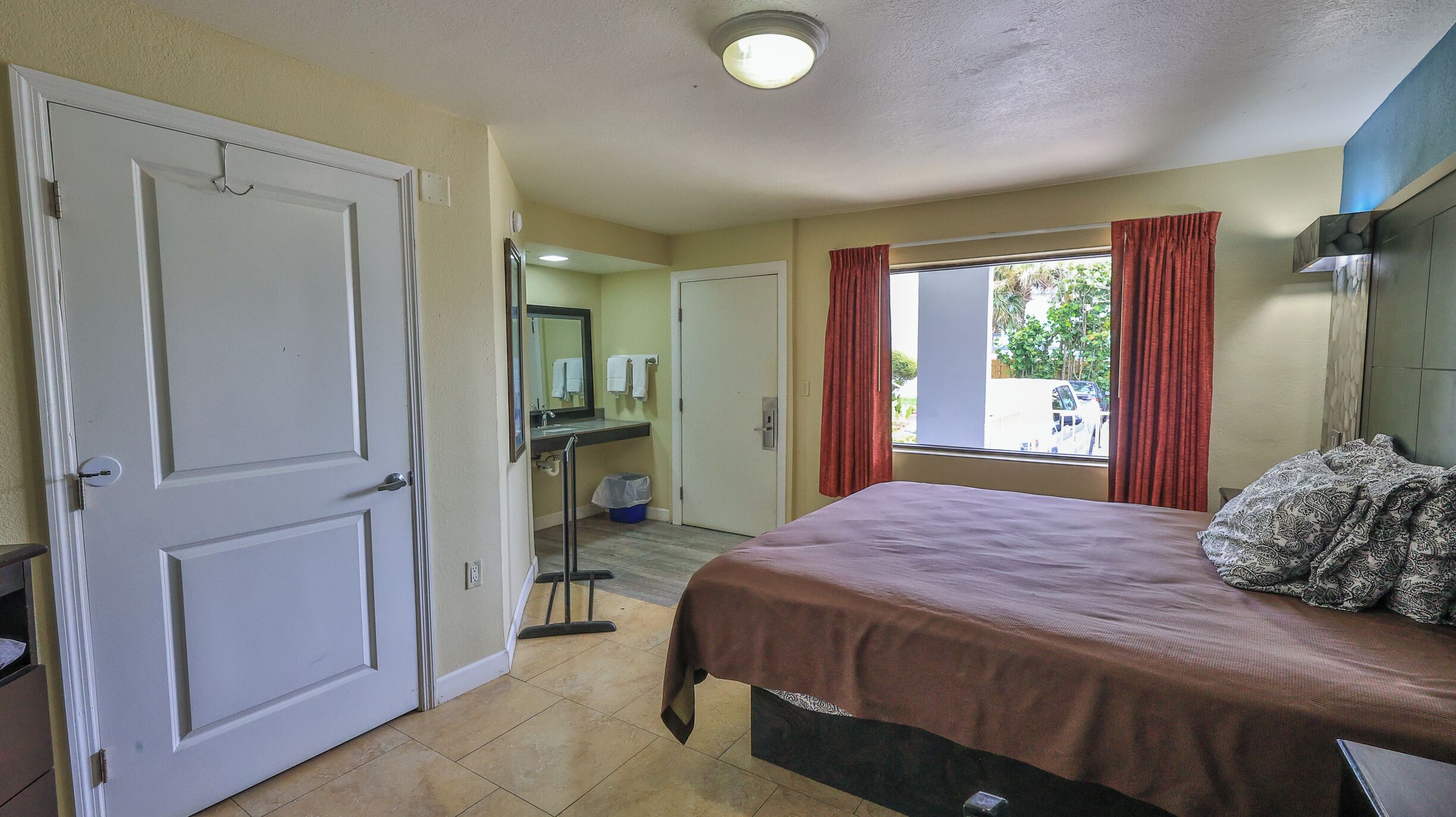 Une chambre d’hôtel avec un lit, une fenêtre avec des rideaux, une salle de bain avec un miroir et une porte.