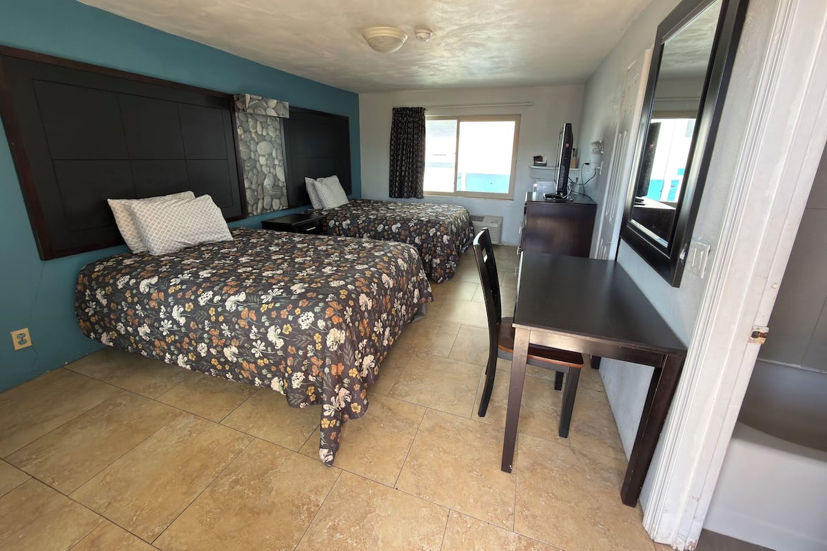 Una habitación con dos camas, un escritorio, una silla, un ventanal y un espejo.