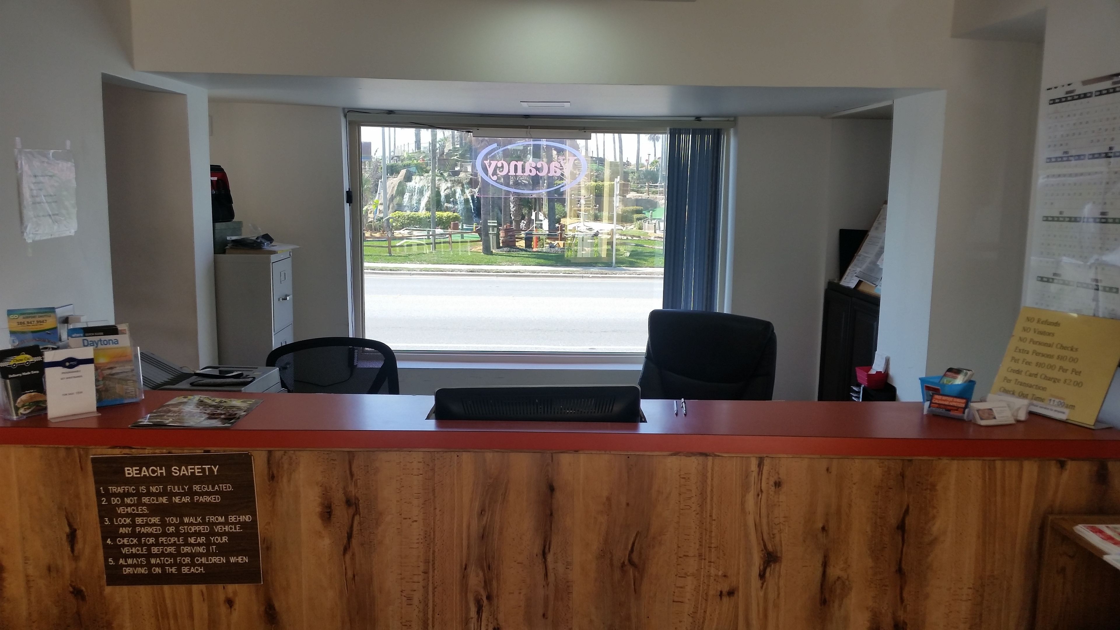 A reception desk with a wooden panel, a computer, and a chair. Outside, there's a view of a parking lot and a building with a neon sign.