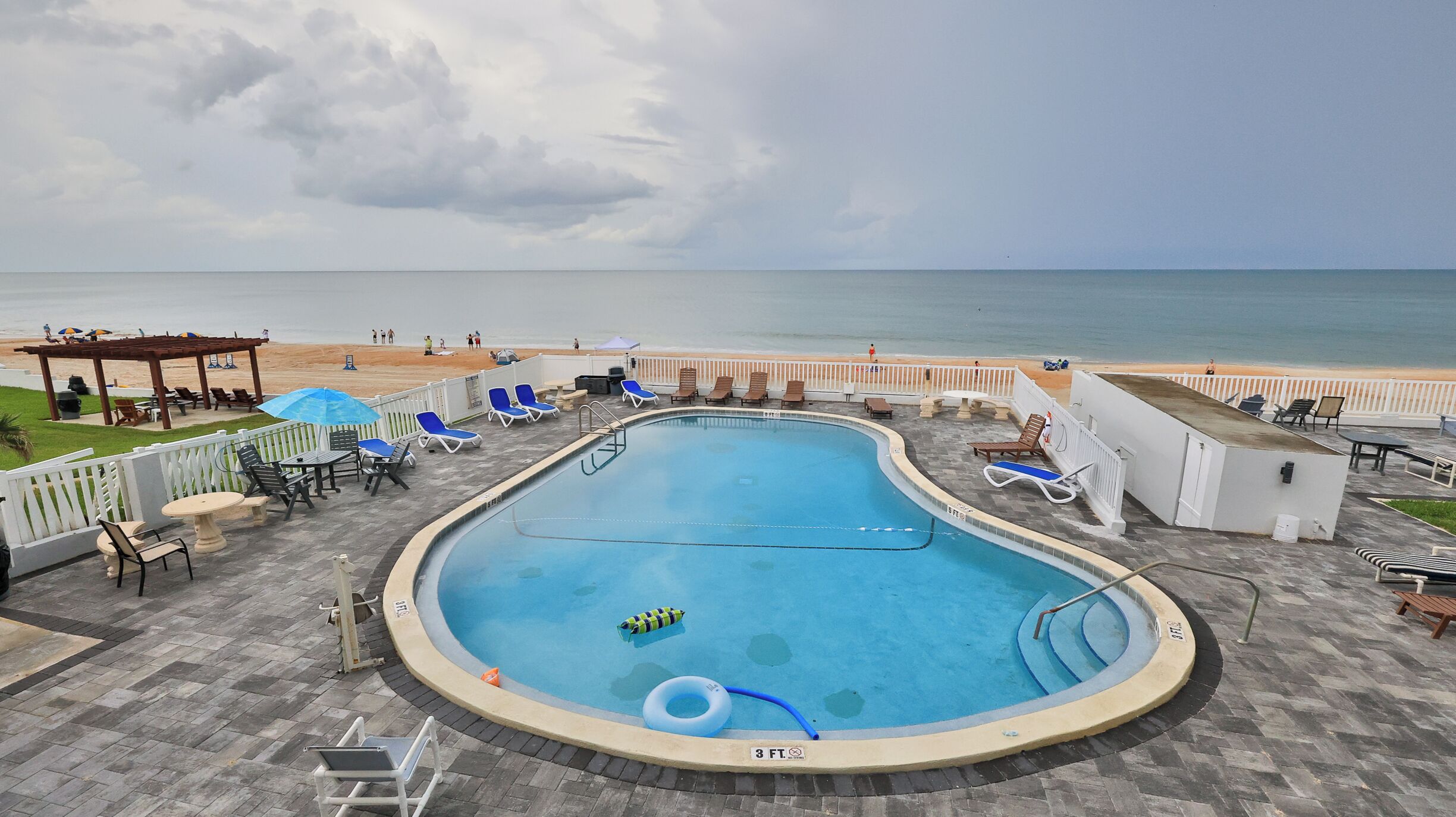 A swimming pool with a view of the beach, lounge chairs, and a covered area with umbrellas.
