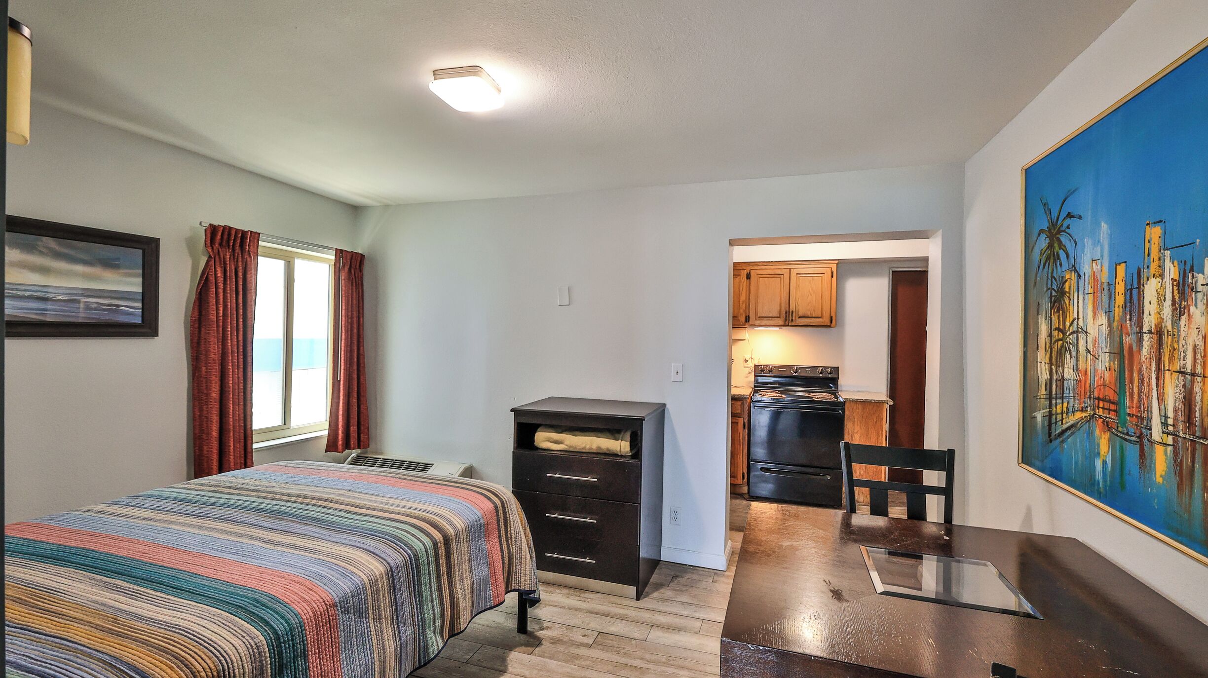 A compact bedroom with a bed, a dresser, and a painting. A kitchen is visible through an open doorway.