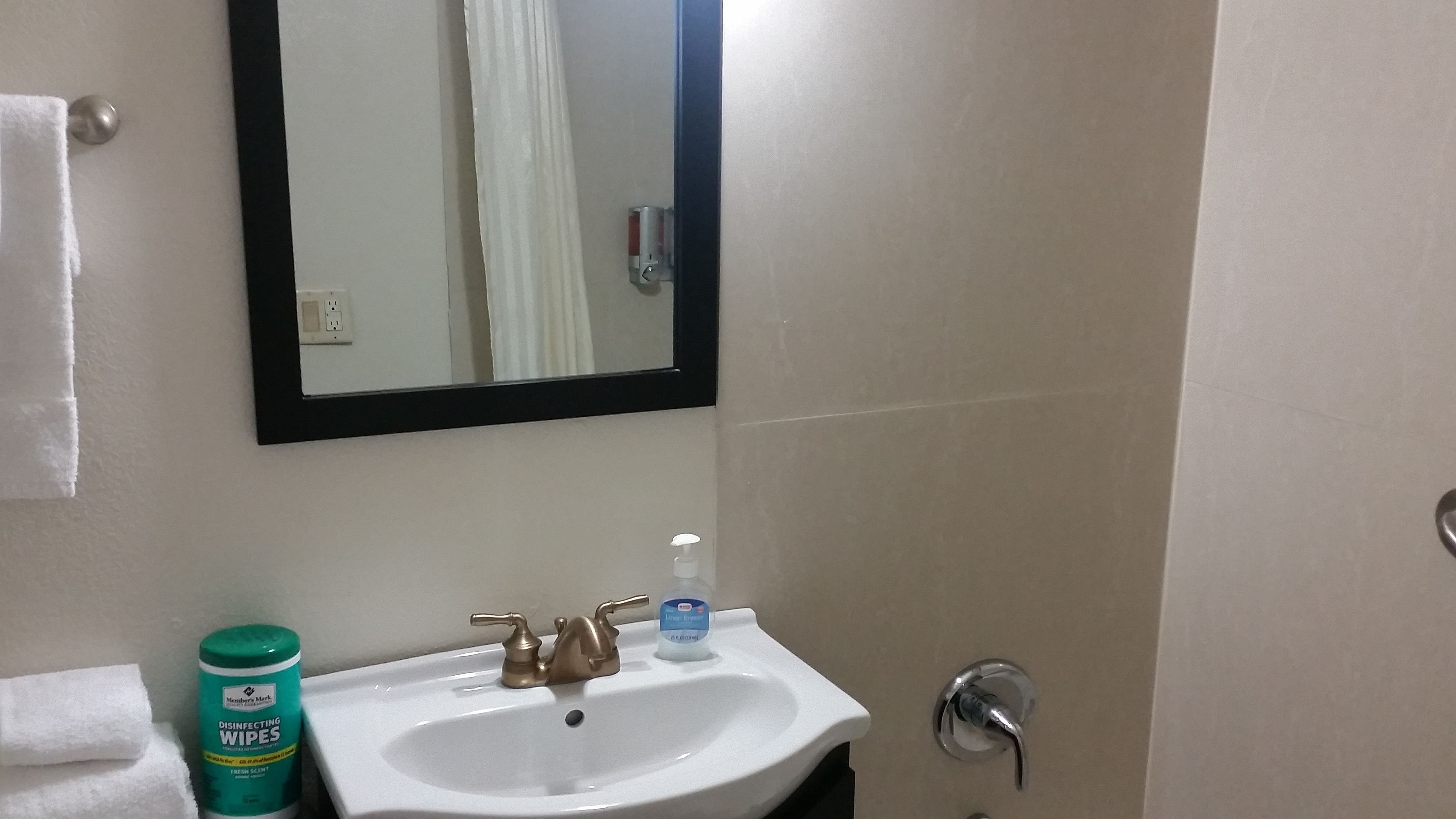 Un baño con un lavamanos blanco, un espejo, una toalla y una botella de toallitas desinfectantes.