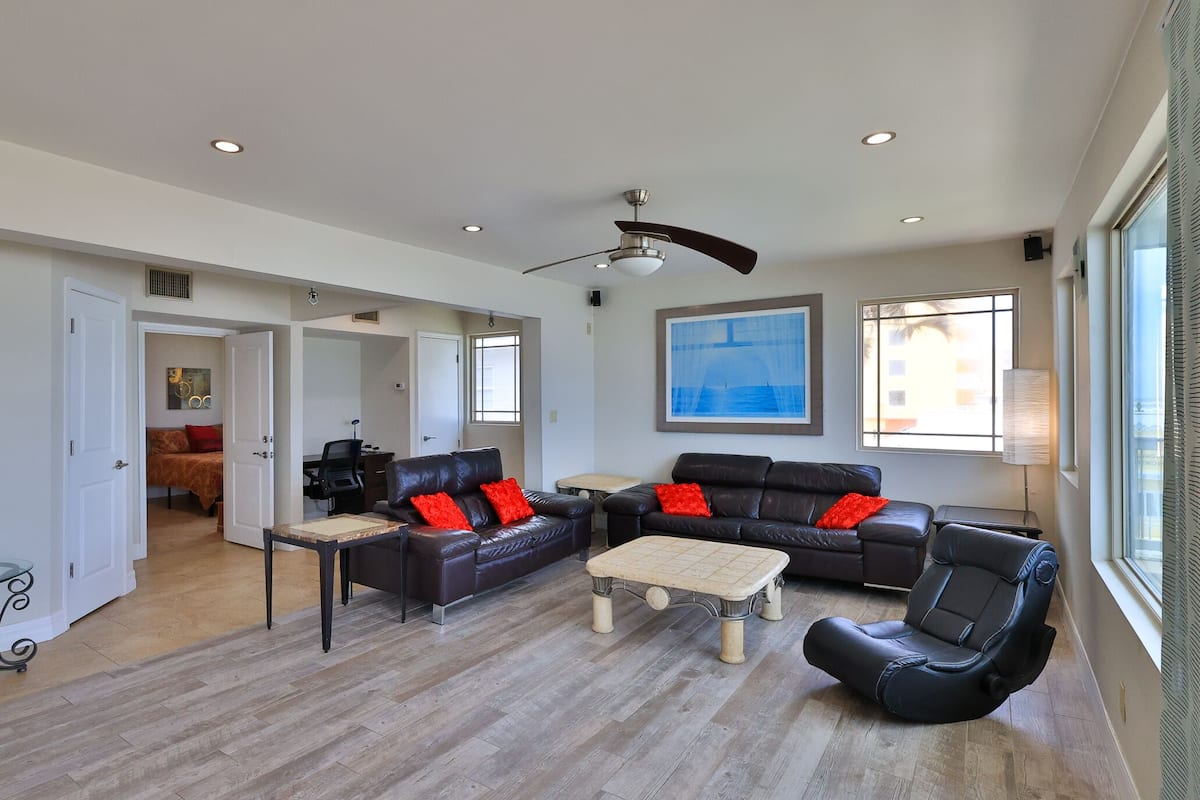 Amplia sala de estar con un sofá grande de cuero negro, una mesa de centro y una mesa de comedor. Hay un ventilador de techo, un cuadro en la pared y una ventana con cortinas.