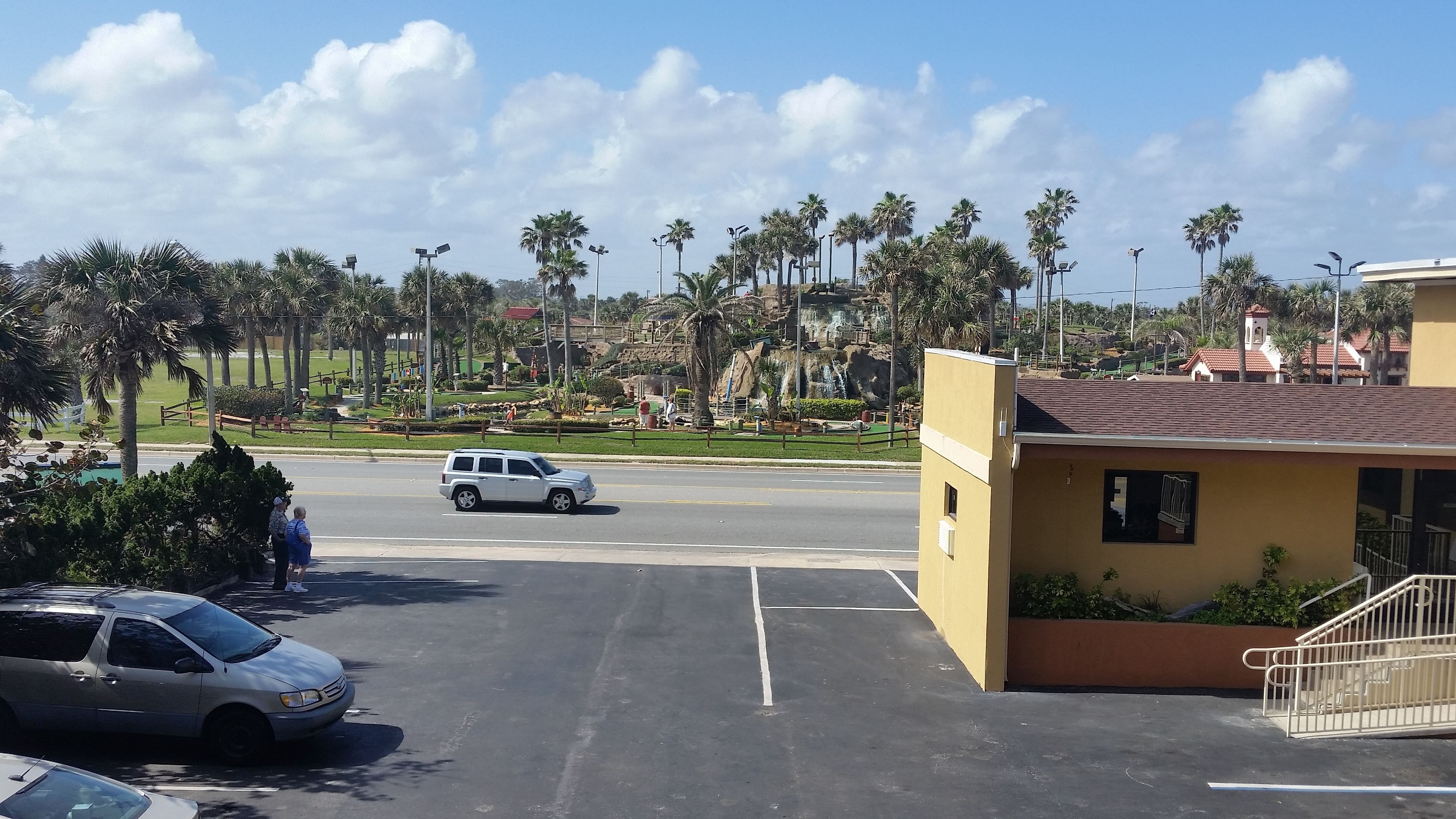 A parking lot with a white SUV, a yellow building, and palm trees in the background.