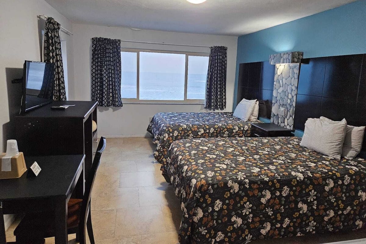 Una habitación de hotel con dos camas, un escritorio con computadora, una ventana con cortinas y decoración de pared de piedra.