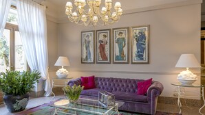 A living room with a purple sofa, a glass coffee table, a chandelier, framed artwork on the wall, and a potted plant. - Grand Hotel Tettuccio (Montecatini Terme)