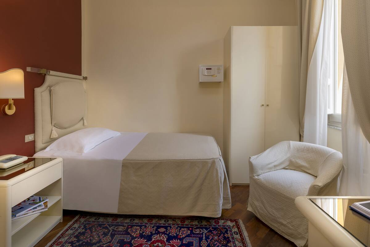 Ein Hotelzimmer mit Bett, Sessel, Nachttisch, Lampe, Thermostat und einem Fenster mit Vorhängen.