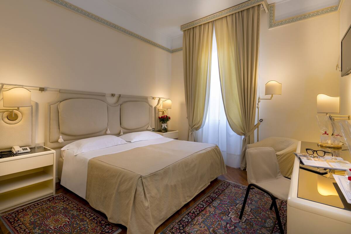 Ein Hotelzimmer mit einem großen Bett, Nachttischen, einem Schreibtisch mit Stuhl, einem Fenster mit Vorhängen und einem Fernseher.