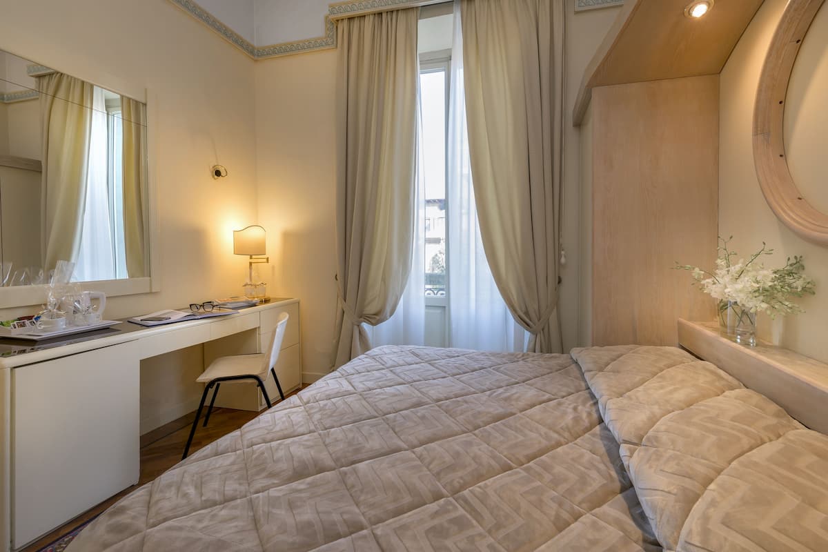 Ein Schlafzimmer mit Bett, Schreibtisch, Stuhl, Spiegel, Lampe und einem Fenster mit Vorhängen.