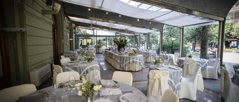 An outdoor dining area with round tables set for a meal, covered by a translucent roof, and surrounded by white chairs.