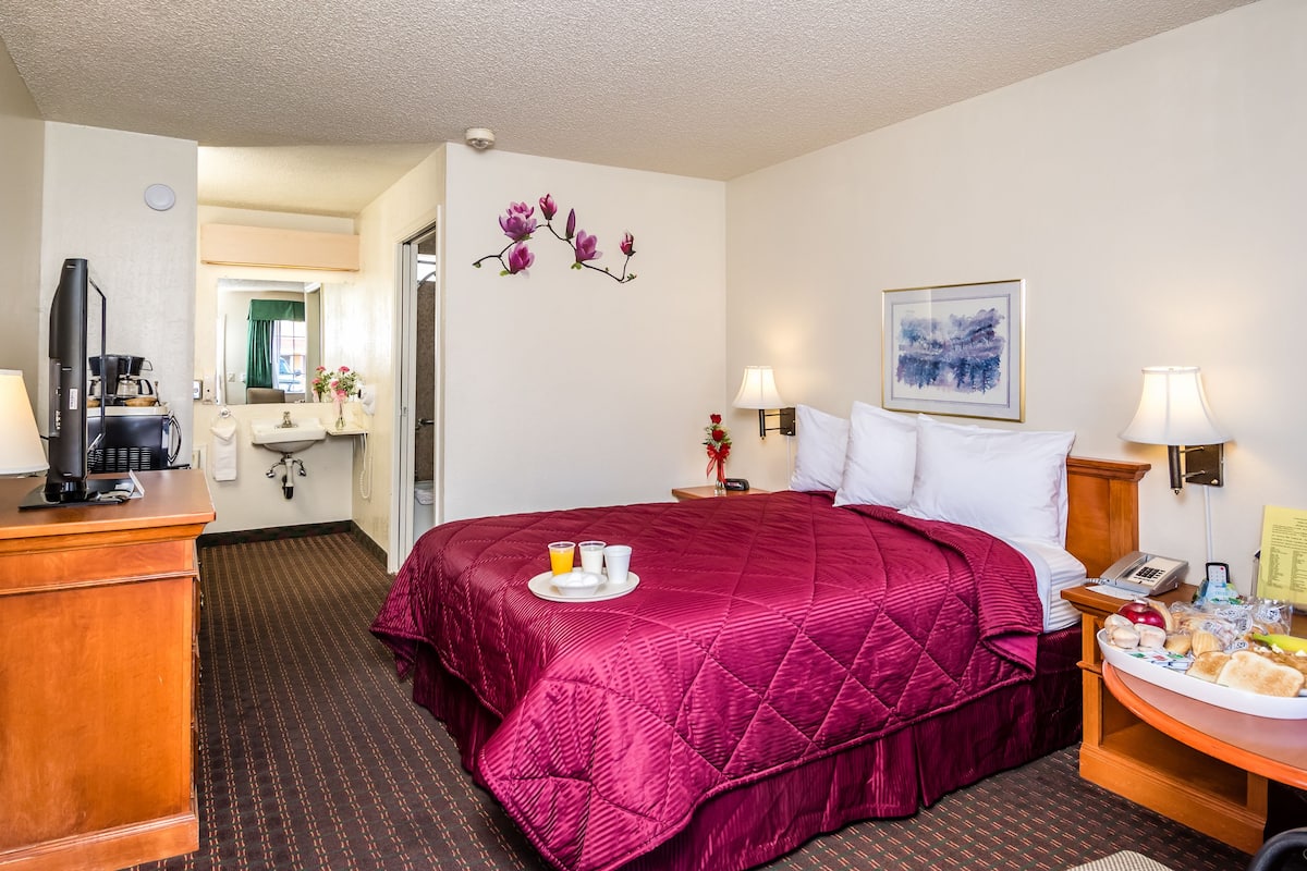 A hotel room with a large bed, a desk with a coffee maker, a bedside table with a lamp, and a painting on the wall.