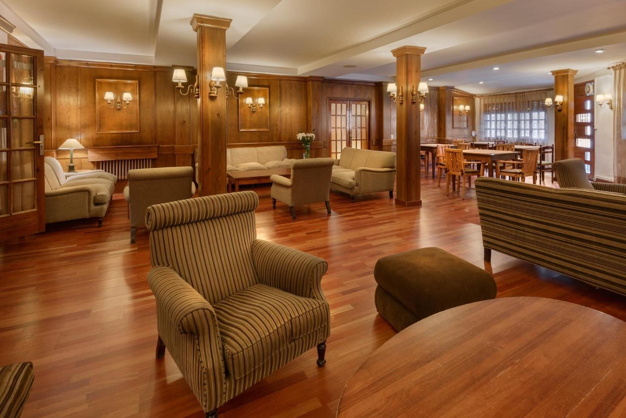 A spacious lobby with wooden paneling, multiple seating areas including sofas and armchairs, and a dining area with wooden tables and chairs.