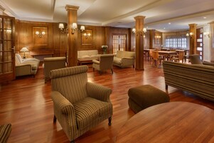 A spacious lobby with wooden paneling, multiple seating areas including sofas and armchairs, and a dining area with wooden tables and chairs.