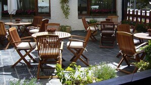 Een terras buiten met houten stoelen en tafels, witte parasols en plantenbakken.