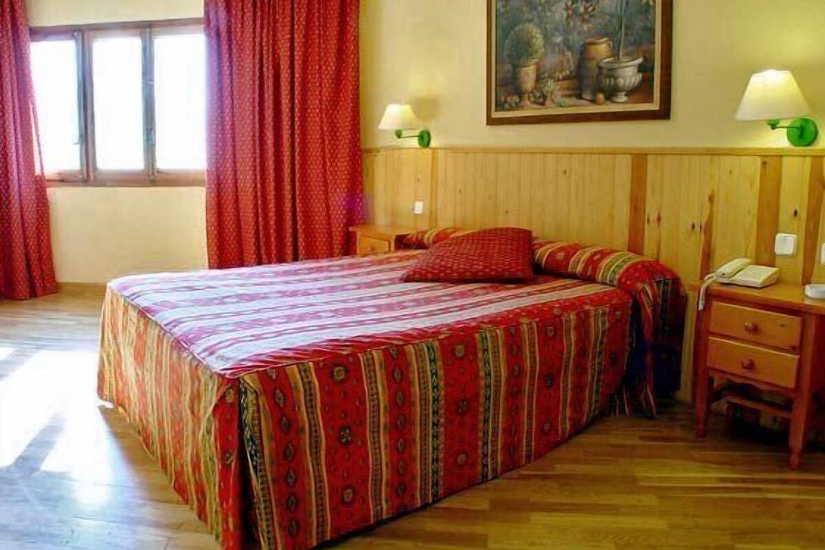 Een kamer met een groot bed, een houten hoofdbord, twee nachtkastjes met lampen, een schilderij aan de muur en een raam met gordijnen.
