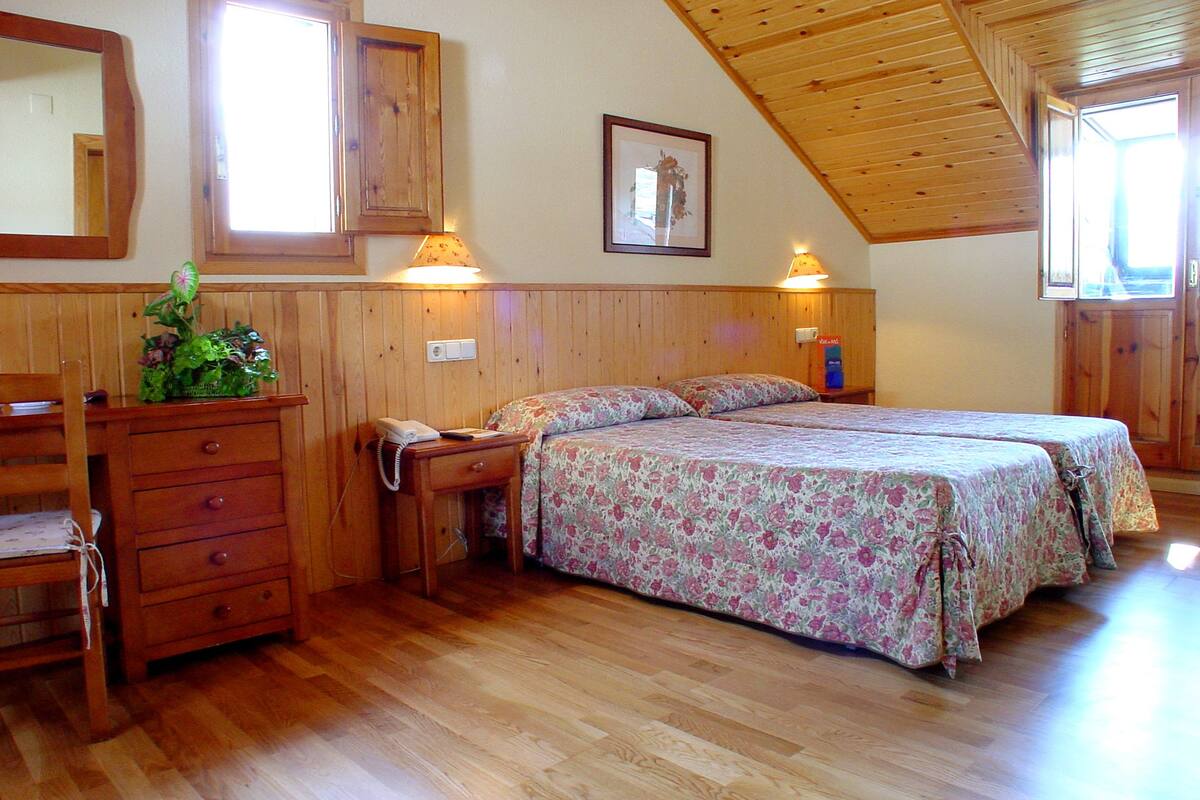Een slaapkamer met houten wanden, een bed met bloemenbeddengoed, een dressoir, een bedlampje en een ingelijste foto aan de muur.
