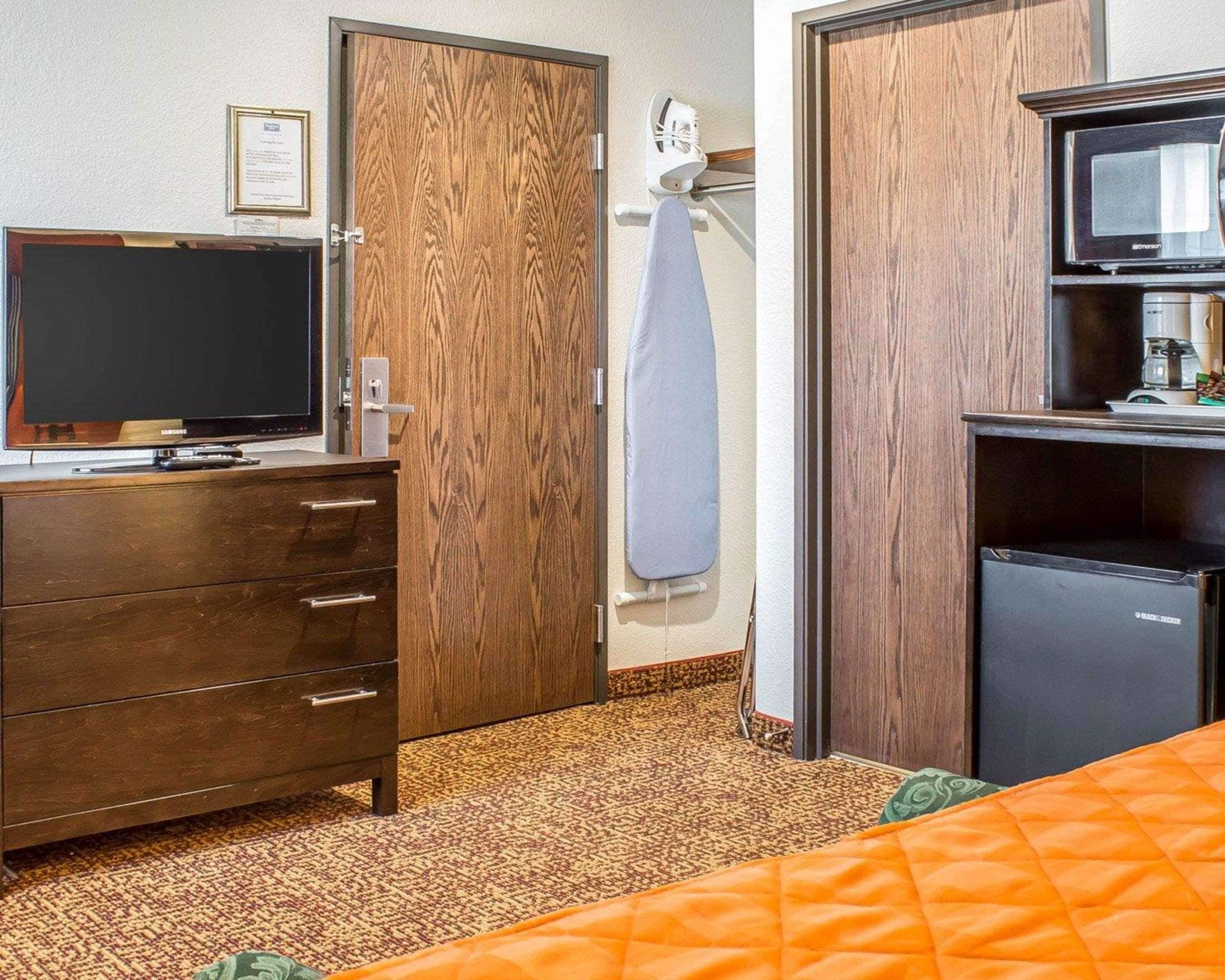 Quarto de hotel com guarda-roupa de madeira, televisão em penteadeira, minibar e cama com colcha laranja.