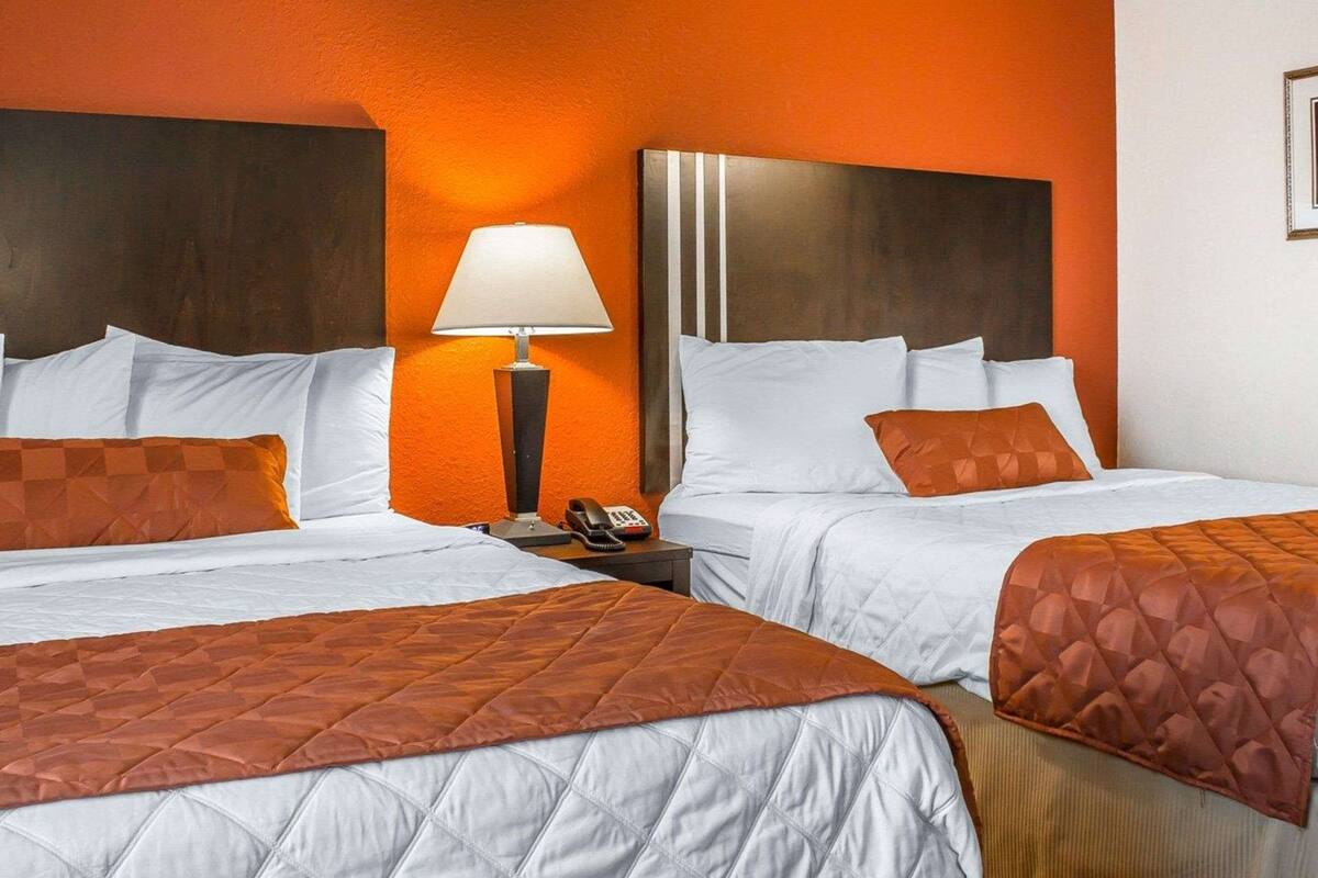 A hotel room with two beds, each with white bedding and orange quilted covers. There is a nightstand with a lamp and a phone between the beds. The room has orange walls and a dark headboard.