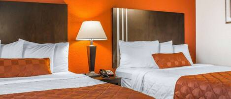 Habitación de hotel con dos camas, cada una con ropa de cama blanca y cubrecamas acolchados anaranjados. Entre las camas, hay una mesita de noche con una lámpara y un teléfono. Las paredes son anaranjadas y el cabecero es oscuro.
